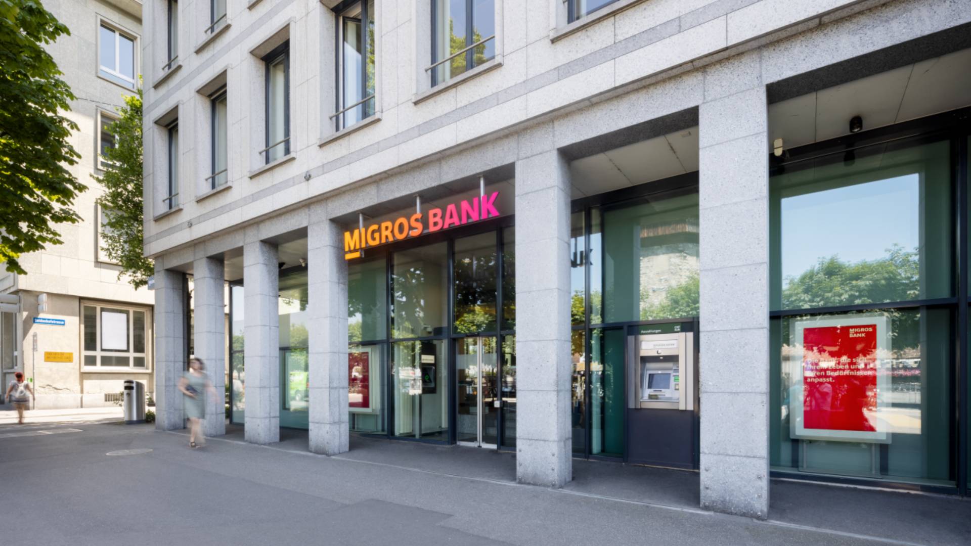 Exterior view of a Migros Bank branch with modern stone facade, large glass fronts, and illuminated logo; pedestrians in front of the entrance and ATM visible, symbolizing banking, customer experience, and customer relationship management with the BSI Customer Suite