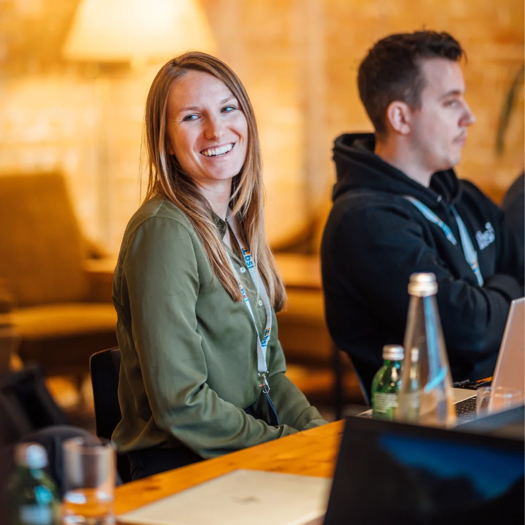 Participants in a workshop or training session sit together in a relaxed setting with laptops and drinks. A woman wearing a name badge smiles at the camera. The scene represents collaborative learning, professional development, and hands-on knowledge sharing with the BSI Academy.