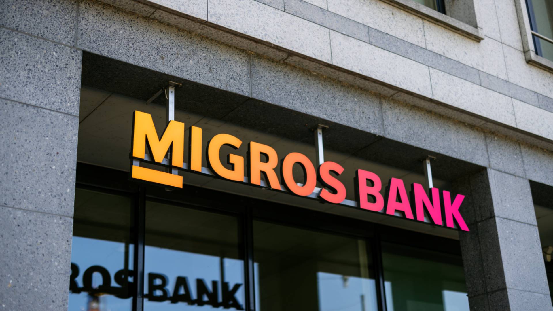 Exterior view of a Migros Bank branch with illuminated logo on a modern building facade; symbol of banking, customer experience, and customer relationship management with the BSI Customer Suite