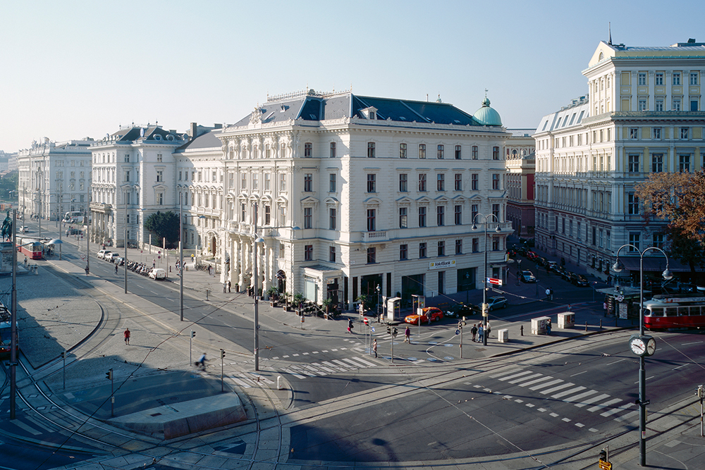Historic street scene in Vienna featuring grand 19th-century buildings, trams, and wide intersections under a clear sky.