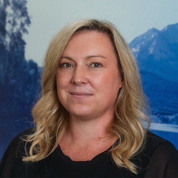 Portrait of a smiling woman with blonde wavy hair in front of a mountain and forest landscape background.