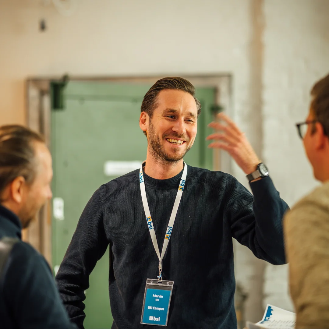 Participants engage in conversation in a relaxed workshop or networking environment. A man wearing a BSI badge gestures while smiling and speaking with other attendees. The scene represents personal knowledge sharing, community, and hands-on professional development with the BSI Academy.