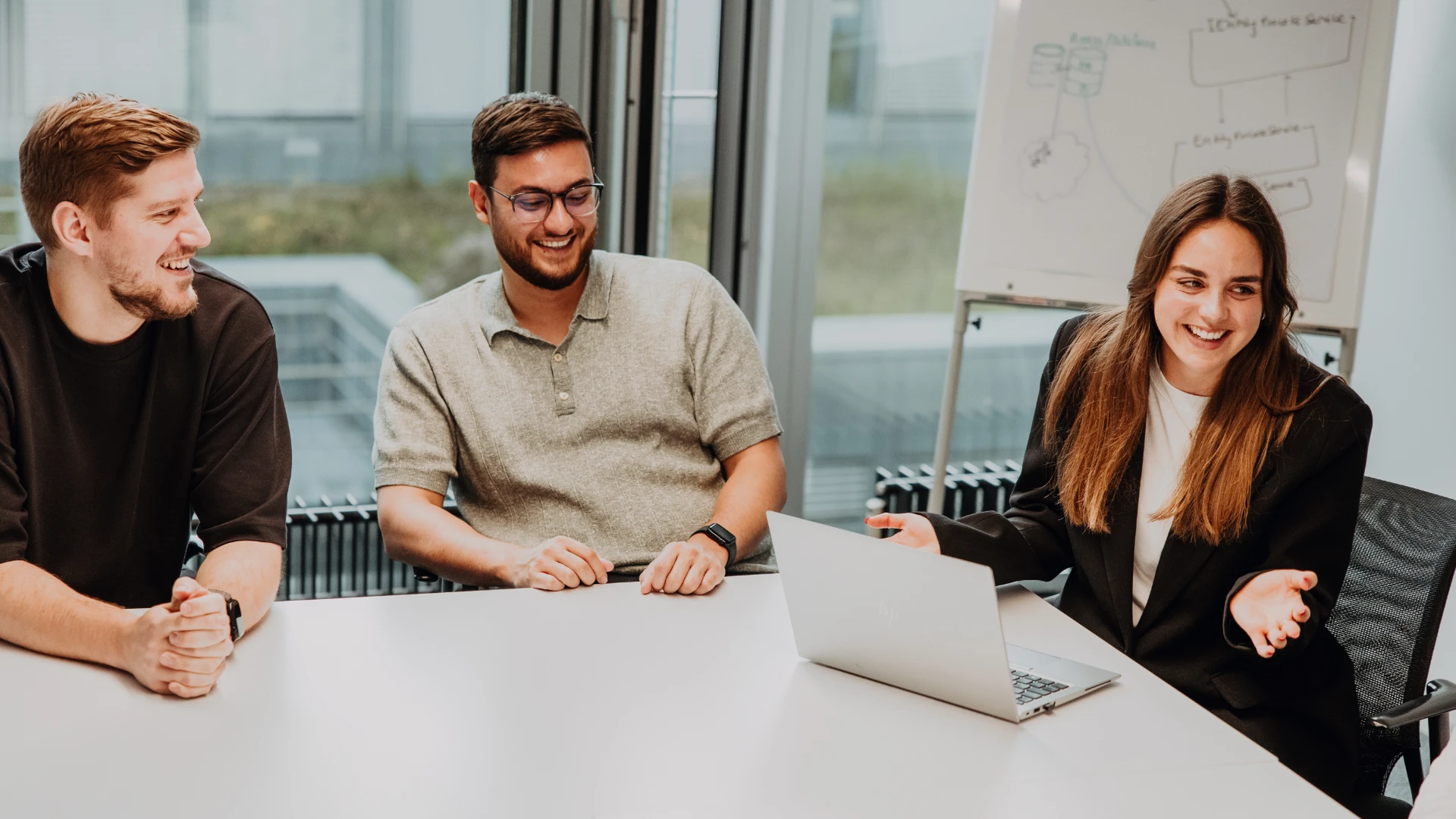 Three employees sit together in a meeting room, laughing and discussing ideas around a laptop—an open, supportive team culture and modern work environment at BSI Software as an attractive employer.
