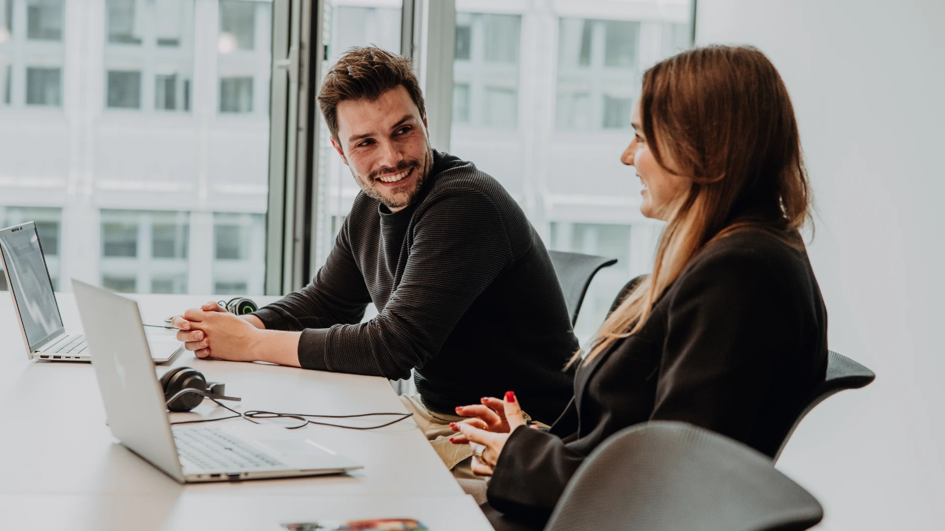 Two employees collaborate on laptops in a modern office, smiling and exchanging ideas, reflecting an open and supportive work environment—an authentic glimpse into the culture and working at BSI Software.