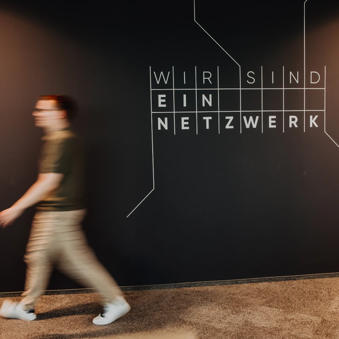 An employee walks past a wall reading “We are a network” — modern office design and a strong sense of connection reflect the open culture and attractive work environment at BSI Software.