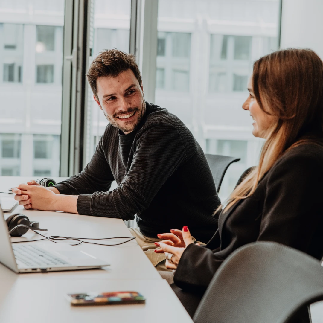 Two employees collaborate on laptops in a modern office, smiling and exchanging ideas, reflecting an open and supportive work environment—an authentic glimpse into the culture and working at BSI Software.