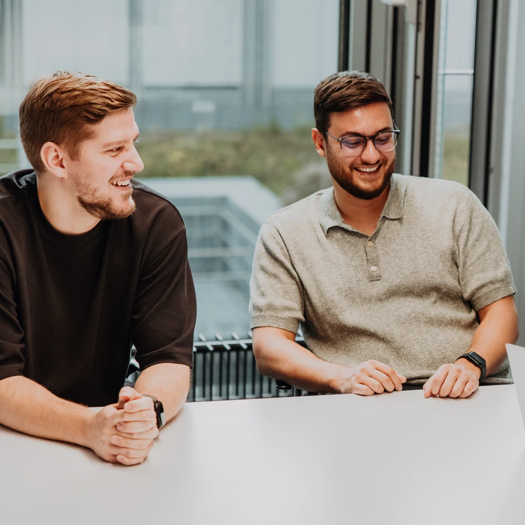 Three employees sit together in a meeting room, laughing and discussing ideas around a laptop—an open, supportive team culture and modern work environment at BSI Software as an attractive employer.